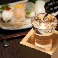 Sake in a box: A glass is set inside a wooden masu cup and is being filled to overflow. Behind it is a plate of sushi.