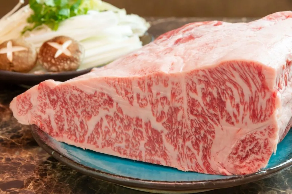 A large piece of beautifully-marbled wagyu sits on a blue plate ready to be cuts into slices. In the background, there is a plate of various vegetables waiting to be cooked. The Japanese system of wagyu grading is extremely strict, with the highest being a5 wagyu.