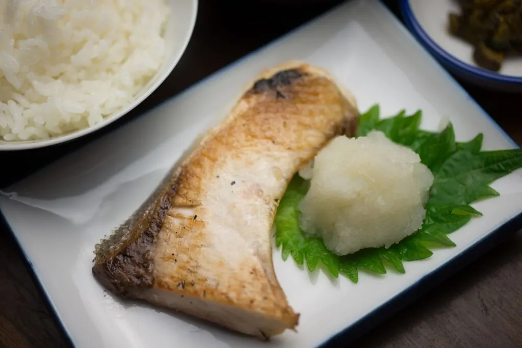 A piece of grilled buri served with grated radish and a shiso leaf. A bowl of white rice can also be partially seen in the top left-hand corner.