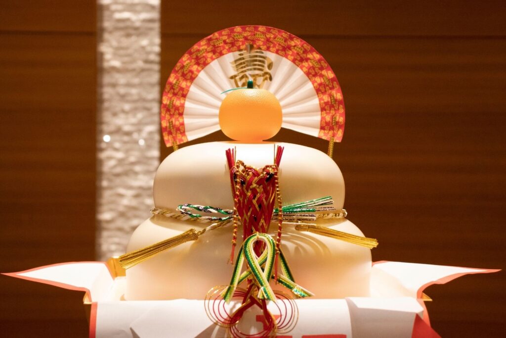 A plastic kagami mochi decoration housing the two mochi rice cakes inside. Atop is a plastic bitter orange and a paper fan.