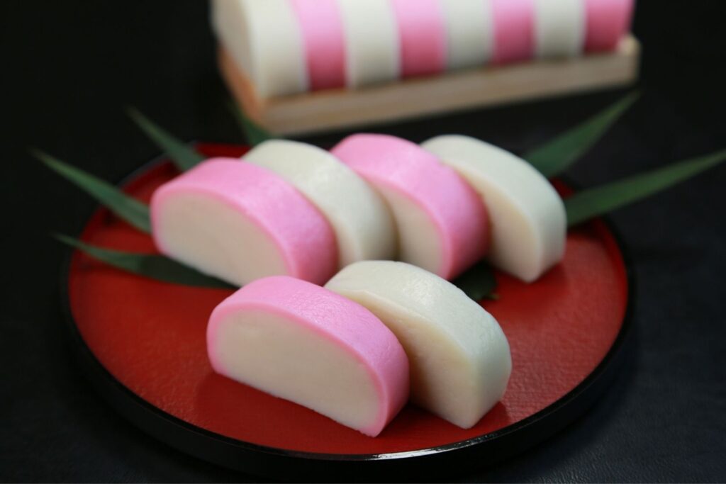 Cut up kohaku kamaboko presented in an alternating color pattern. The pink fish cake (representing red) is pink on the outside but white on the inside; the white one is simply white all over.
