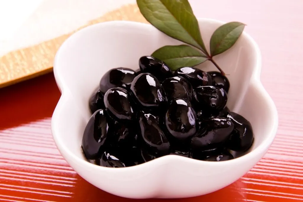 A small white bowl filled with shiny black kuromame beans and a leaf for presentation.