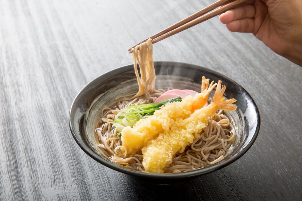 Toshikoshi soba in a dark bowl with kohaku kamaboko fish cake, two tempura shrimp and spring onions, set on a wooden surface. A hand can be seen on the right side of the image holding chopsticks and lifting up some of the soba noodles.