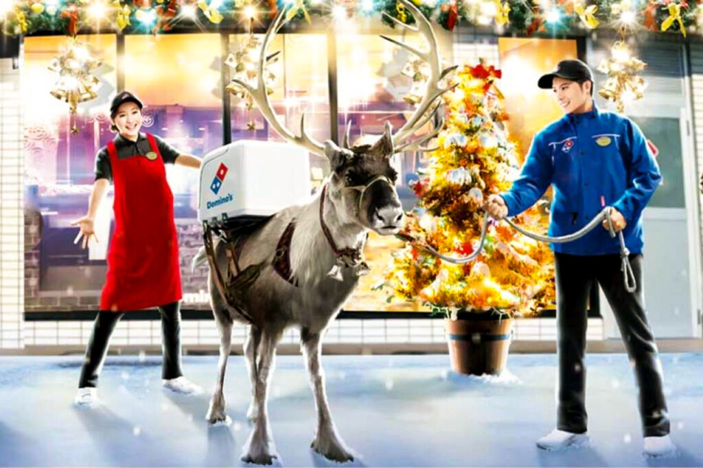 A reindeer with a Domino's delivery container strapped to its back stands outside a festively-decorated Domino's Japan store with a Christmas tree out front. Two staff members, a male staff member in blue holding the reindeer's reins and a female staff member in red with her hands outstretched, look on with delight.