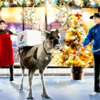 A reindeer with a Domino's delivery container strapped to its back stands outside a festively-decorated Domino's Japan store with a Christmas tree out front. Two staff members, a male staff member in blue holding the reindeer's reins and a female staff member in red with her hands outstretched, look on with delight.