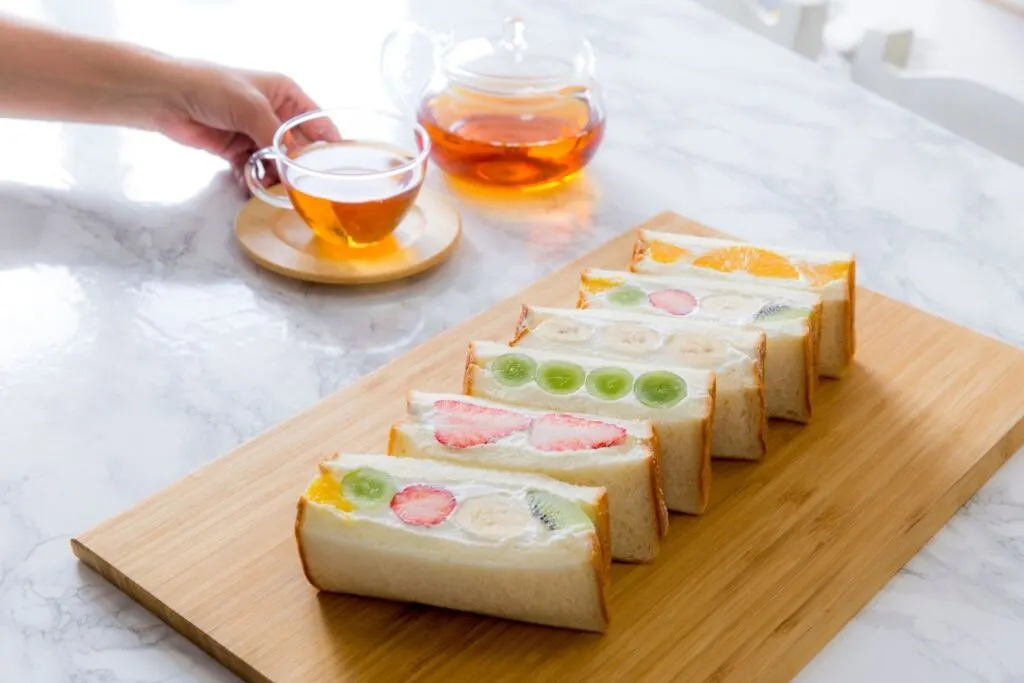 A wooden chopping board with six half Japanese fruit sandwiches on it - one each of orange, banana, green grape and strawberry fillings, and two with all of those fruits plus kiwi. Behind the board is a clear glass tea pot and matching tea cup filled with reddish-black tea on a wooden coaster.