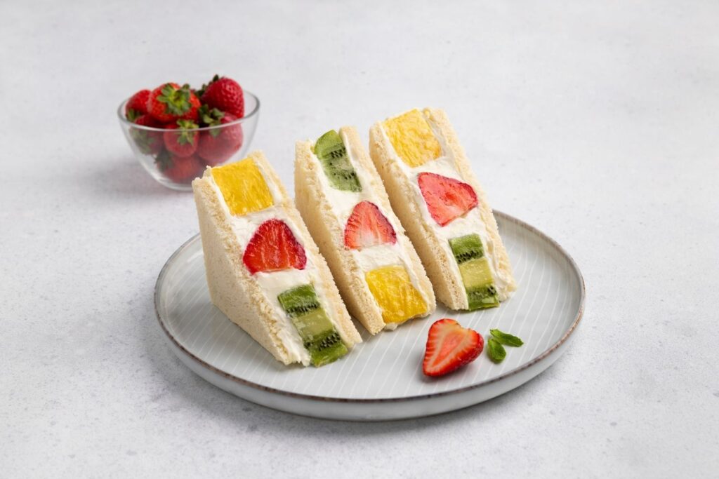 Three half slices of Japanese fruit sandwiches with pineapple, strawberry and kiwi on a plate garnished with one half strawberry. Behind the plate is a small clear glass bowl filled with whole strawberries.