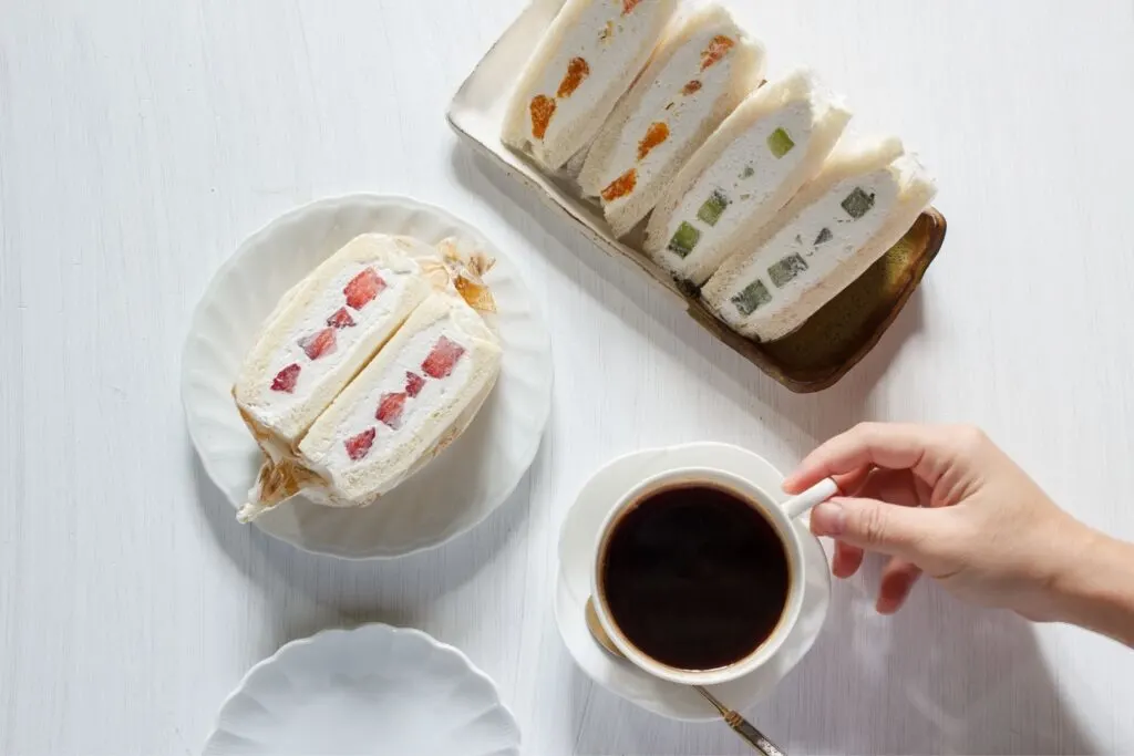 Two diagonally-cut Japanese fruit sandwiches, one with orange and the other with kiwi fruit, sit on a white rectangular plate. In front of that is a white round plate with a rectangular-cut fruit sando with strawberry. To the right of the plate, a hand can be seen picking up a cup of black coffee off a saucer.