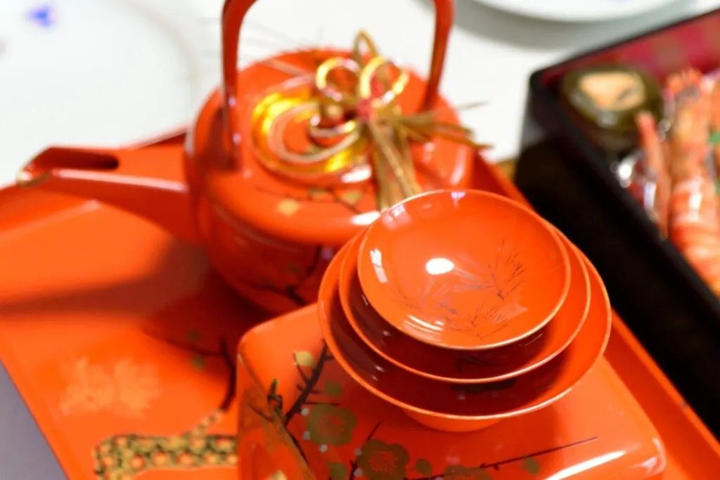 Ready to drink toso: A decorative vermillion new year sake teapot with three stacked matching sake cups in front of it. To the right of the image, the corner of a box of osechi New Year cuisine can be seen.