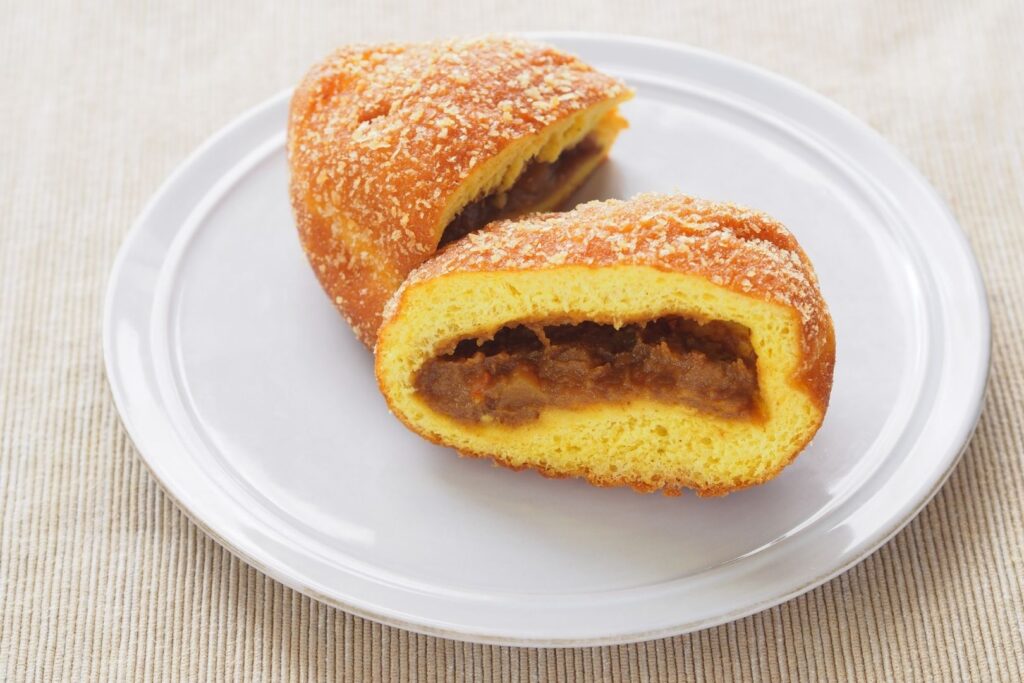 Surprising Japanese Food Names: A deep-fried curry pan broken in half to show the curry filling on a white plate with a decorative ring around the outer rim atop a beige surface.