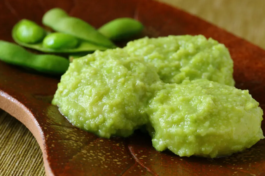 Three large dollops of green zunda paste on a curved wooden plate with some edamame, from which it is made, behind it on the same plate.