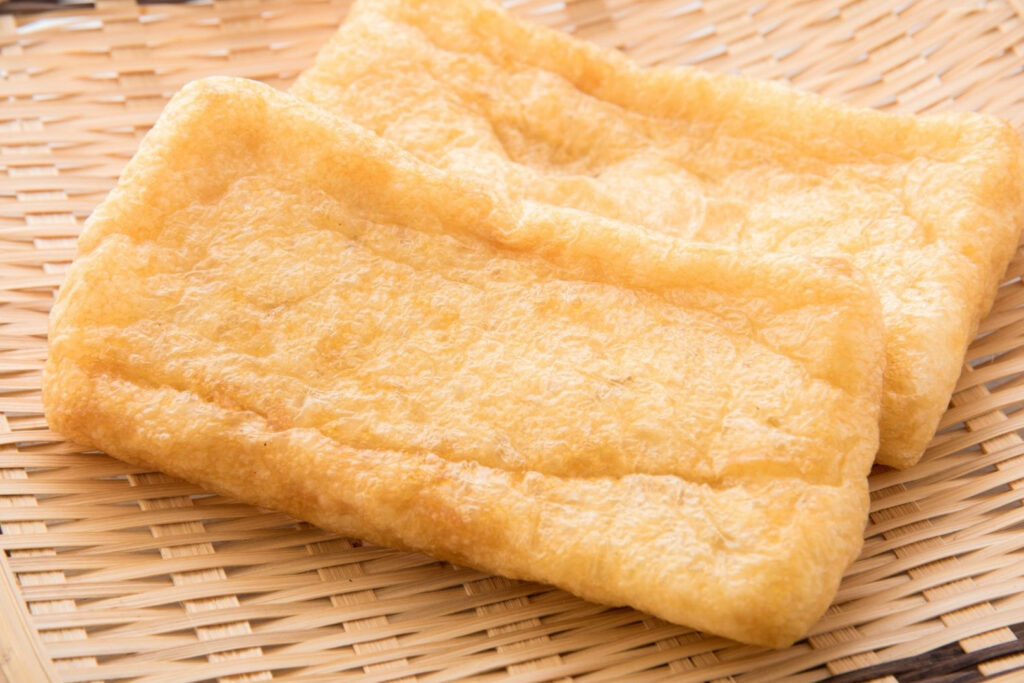 Two pieces of golden-colored deep-fried aburaage sit on a woven bamboo mat.