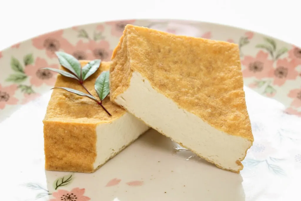 A piece of golden-colored deep-fried atsuage sliced in half diagonally to show the white flesh of the tofu on the inside with a leaf sprig on top. It sits on a decorative plate with pink flowers and green foliage concentrated around the rim. 
