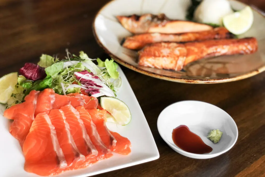 A white square plate with slithers of raw ginzake (branded salmon) served with salad greens and two wedges of lime. Next to it is a small round dish with soy sauce and wasabi, and to the back is three pieces of cooked salmon with grated daikon radish and another wedge of lime.