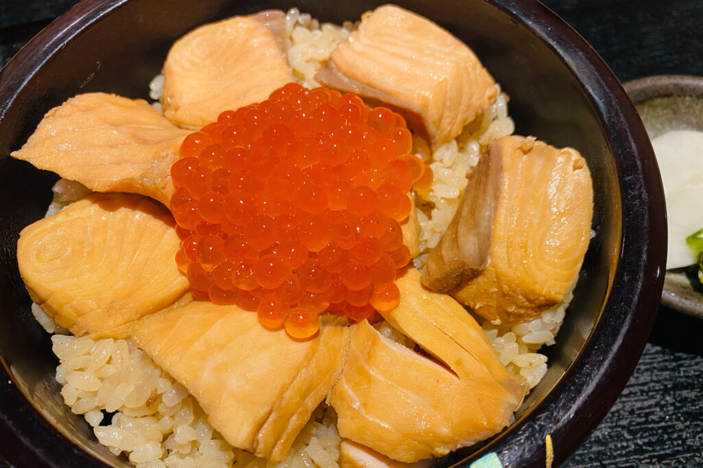 A close-up of a dark wooden bowl filled with seasoned rice along with salmon and its roe on top (a dish known as Harako Meshi in Miyagi Prefecture).