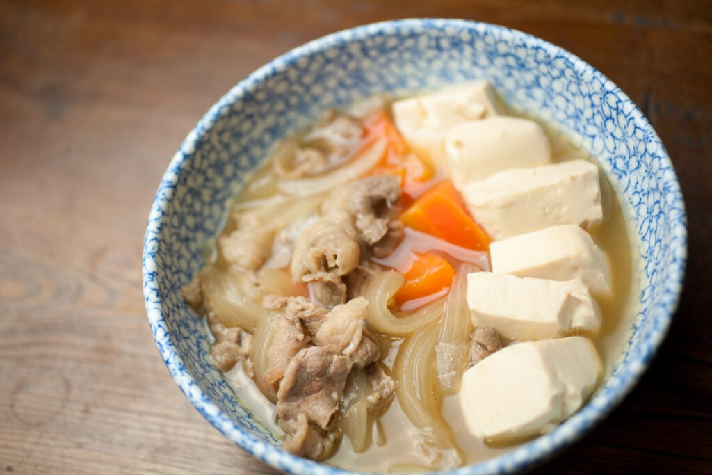 A blue and white dotted ceramic bowl filled with niku-dofu stew. Tofu, meat, carrot and onion can be seen in the broth. The bowl is sitting on a natural wooden surface.