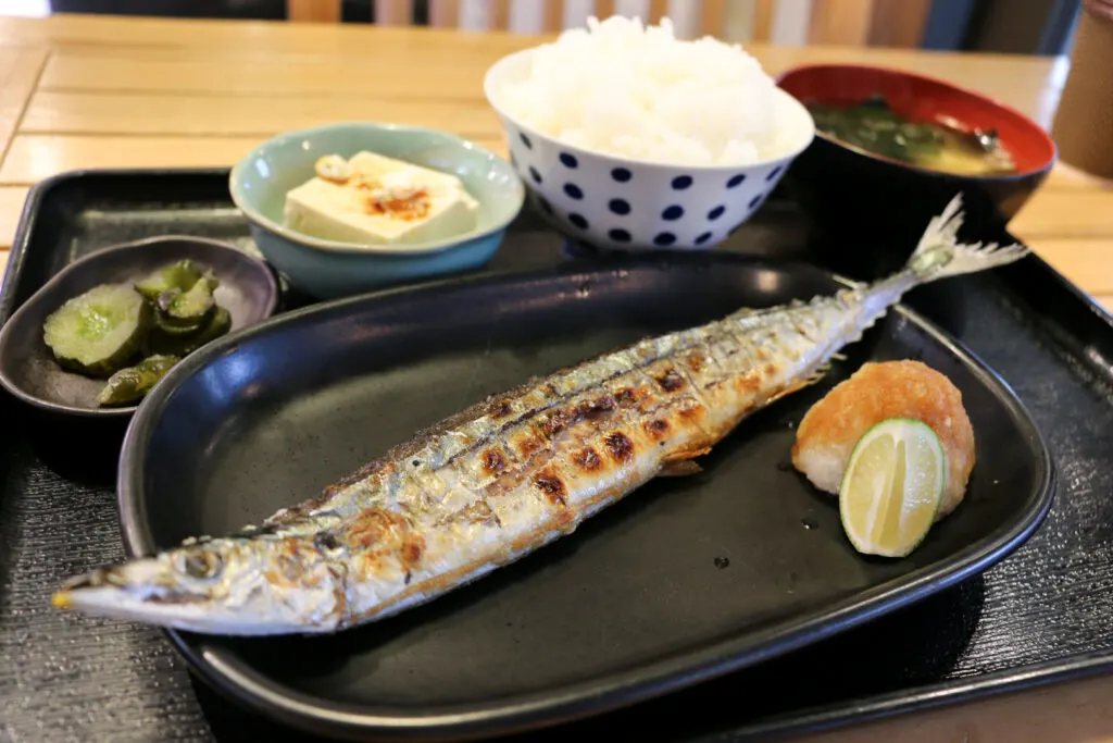 Miyagi Prefecture food: A whole grilled sanma (Pacific saury) fish with a serving of daikon radish and a wedge of lime. Behind the plate are several other side dishes, namely pickles, tofu, a bowl of white rice and a bowl of miso soup. All the dishes are on a black tray, which is sitting on a light-colored wooden countertop.