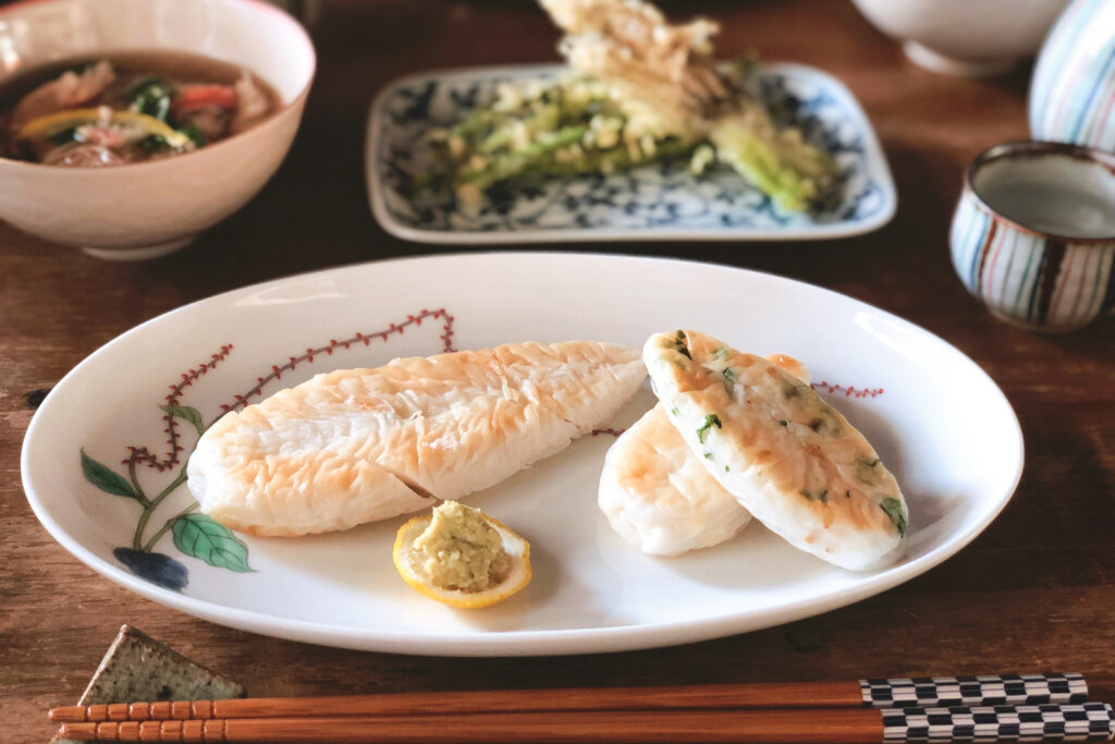 Three pieces of sasa kamaboko arranged on a white plate with an accompanying paste to eat with it. Chopsticks sit on a ceramic chopstick rest in front of the plate, and behind it various other Japanese dishes can be seen on the dark wooden table.  