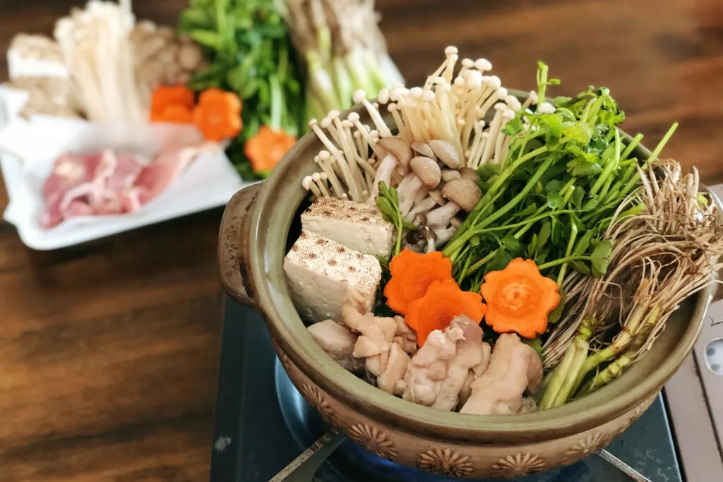 A ceramic nabe pot on a portable gas stove filled with seri nabe ingredients ready to be cooked. This includes of course seri and its roots, as well as mushroom, carrot, tofu and chicken. A plate with additional ingredients can be seen in the background.