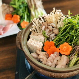 A ceramic nabe pot on a portable gas stove filled with seri nabe ingredients ready to be cooked. This includes of course seri and its roots, as well as mushroom, carrot, tofu and chicken. A plate with additional ingredients can be seen in the background.