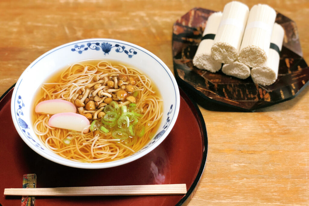 A bowl of hot shiroishi umen noodles in broth with mushrooms, scallions (spring onions) and two slices of kamaboko fish cake on top. The bowl is white with a blue decorative design and sits on a red wooden serving tray with a black rim. There is a pair of wooden disposable chopsticks sitting on a chopstick rest in front of the bowl. To the back right, we can see five rolled up wet hand towels sitting on a black decorative tray.