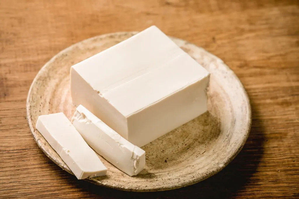 A block of silken tofu with two slices cut off on an earthy-colored ceramic plate upon a wooden surface.