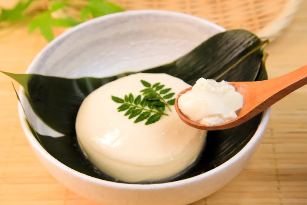 Soft tofu in a rounded shape with two leaf sprigs on top and sitting on a larger flat leaf in a ceramic bowl. A small wooden spoon can be seen coming into the picture from the right with some of the tofu on it (from another piece of tofu).