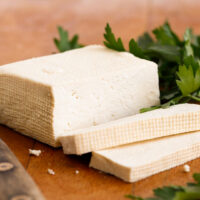 A chunk of firm tofu with two slices cut off on a wooden board. The indentation of the muslin cloth used to strain the tofu can be seen on its surface. A knife is resting to the left of the tofu and some chopped up coriander (cilantro) to the right.