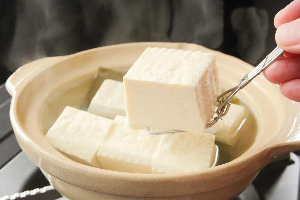 Big chunks of tofu, one being held up for the camera on a small metal strainer from the right-hand side, sit inside a cream-colored nabe hot pot in a dashi broth.