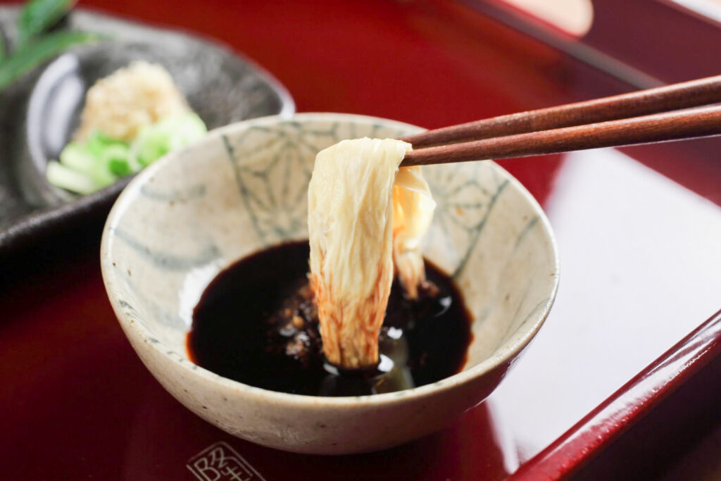 Yuba being dipped into soy sauce with chopsticks from the right-hand side. There is another dish sitting on the red-lacquered table to the back left.