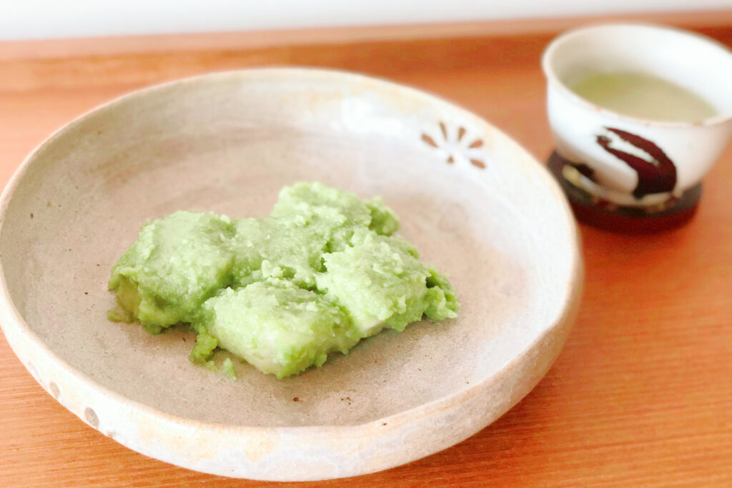 A serving of zunda mochi in a small and shallow light-colored ceramic bowl with a cup of Japanese-style tea to the right.