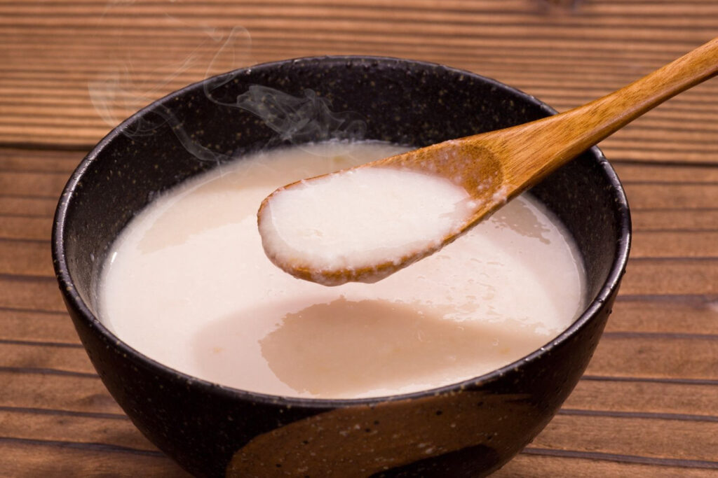 A small black ceramic bowl with flecks of beige is filled with thick, off-white amazake. A natural-colored wooden spoon is coming from the right-hand side and holding up a spoonful of the liquid. The bowl sits on a medium-brown wooden surface.  