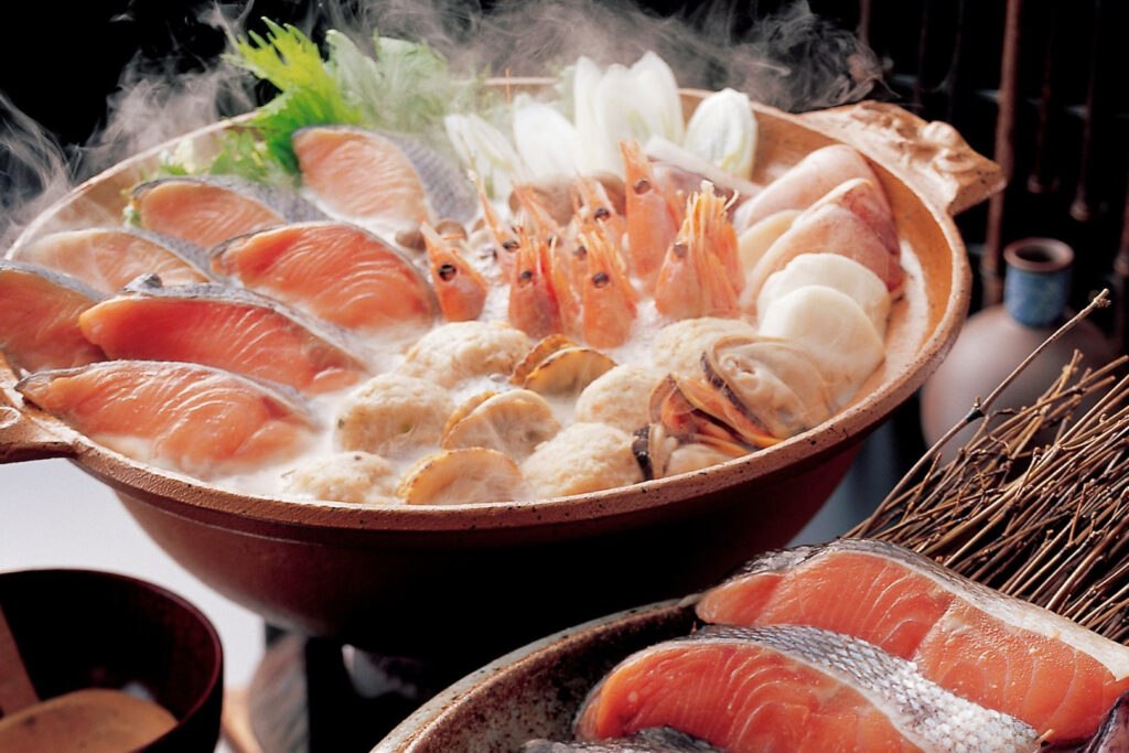 A large brown donabe hot pot filled with Ishikari Nabe ingredients in broth - including salmon, shrimp and other seafood, plus vegetables. In the foreground (right) is a plate with more uncooked salmon on it. On the left is an empty wooden bowl with a wooden ladle in it.