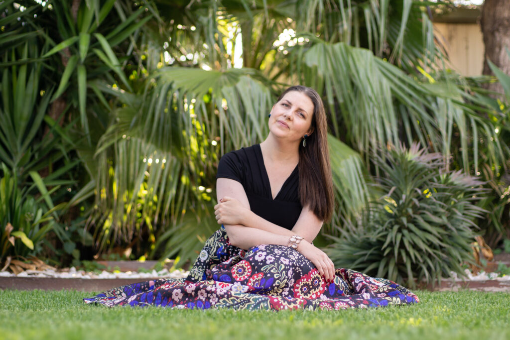 Jessica Korteman, Founder of Japanese Food Guide, sits on the lawn of her Melbourne home with a fan palm and other green leafy plants behind her. She wears a long patterned skirt with royal blue as the dominant color and a black v-neck top with fanned capped sleeves. She has long, brown hair and her head is slightly tilted towards the camera.