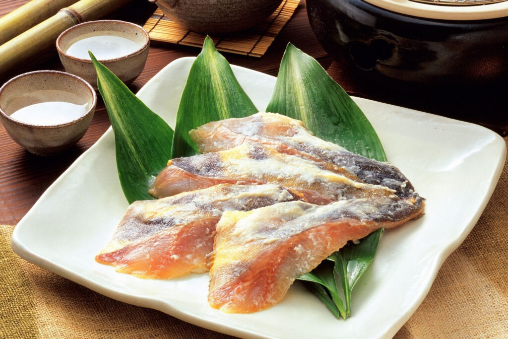 Fish kasuzuke: Four pieces of fish covered in a sake kasu marinade atop three dark green leaves on a large white square plate.