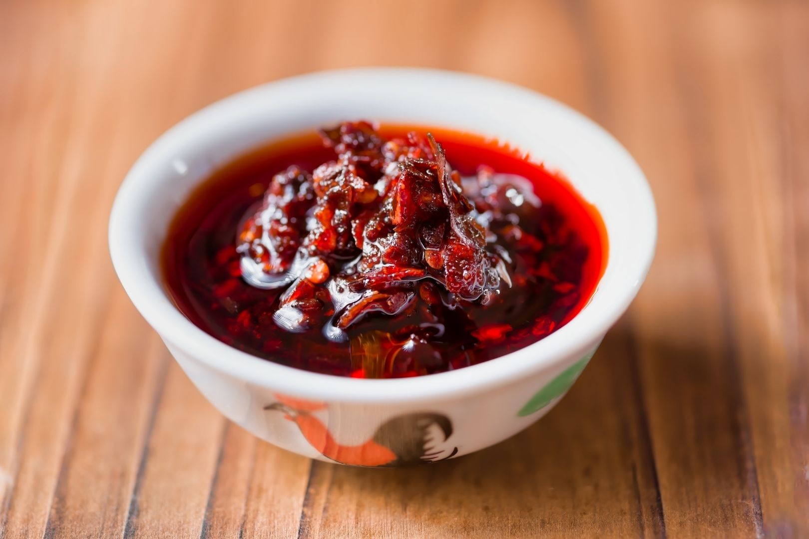 Spicy Japanese condiments: A small white dish with an orange and brown rooster on the front filled with deep red rayu oil. Lots of chopped red chilis can be seen in the oil and are piled up in the center making a peak above the oil. The bowl sits on a natural wooden tabletop.