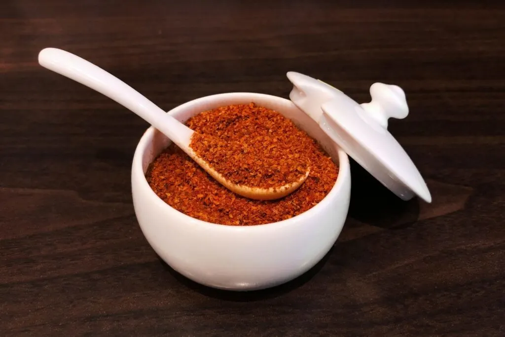 Spicy Japanese condiments: A round white dish with the lid resting up against its side is filled with an earthy red mixture of shichimi togarashi seven spice mix. A white spoon is resting in the dish with a spoonful of the mixture on it.