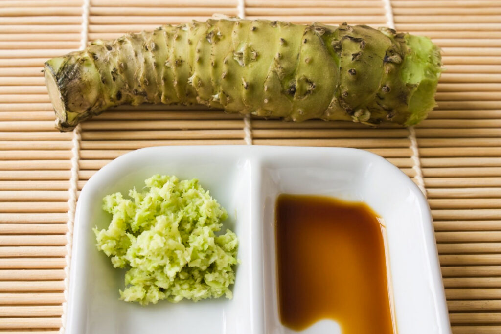 Spicy Japanese condiments: A wasabi rhizome at the back with a white two-compartment dish at the front - grated wasabi on the left side and soy sauce on the right. All items are on a bamboo placement. 