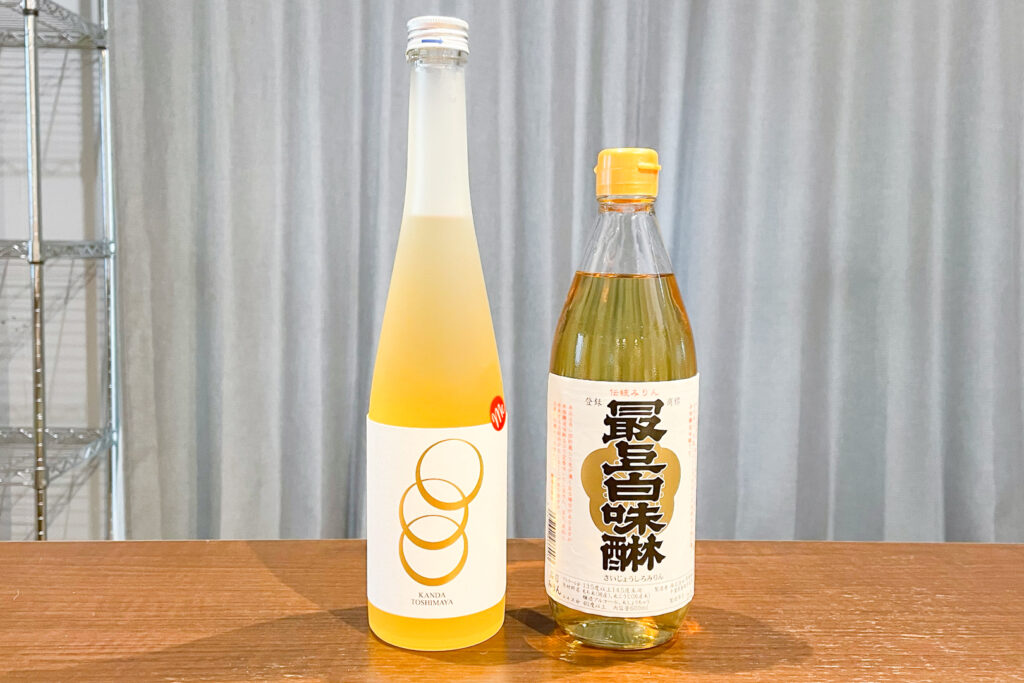 Two bottles of drinking mirin on a wooden table. 'Me' by Toshimaya Shuzo in Tokyo on the left and Saijo Shiromirin by Baba Honten Shuzo in Chiba on the right.