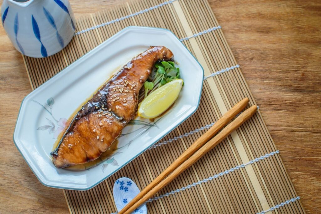 A serving of Japanese amberjack (yellowtail) on a plate with some greens and a slice of lemon. Behind it is a ceramic vessel and in front is a pair of wooden chopsticks on a ceramic chopstick rest. All the items sit on a bamboo mat on a wooden table.  