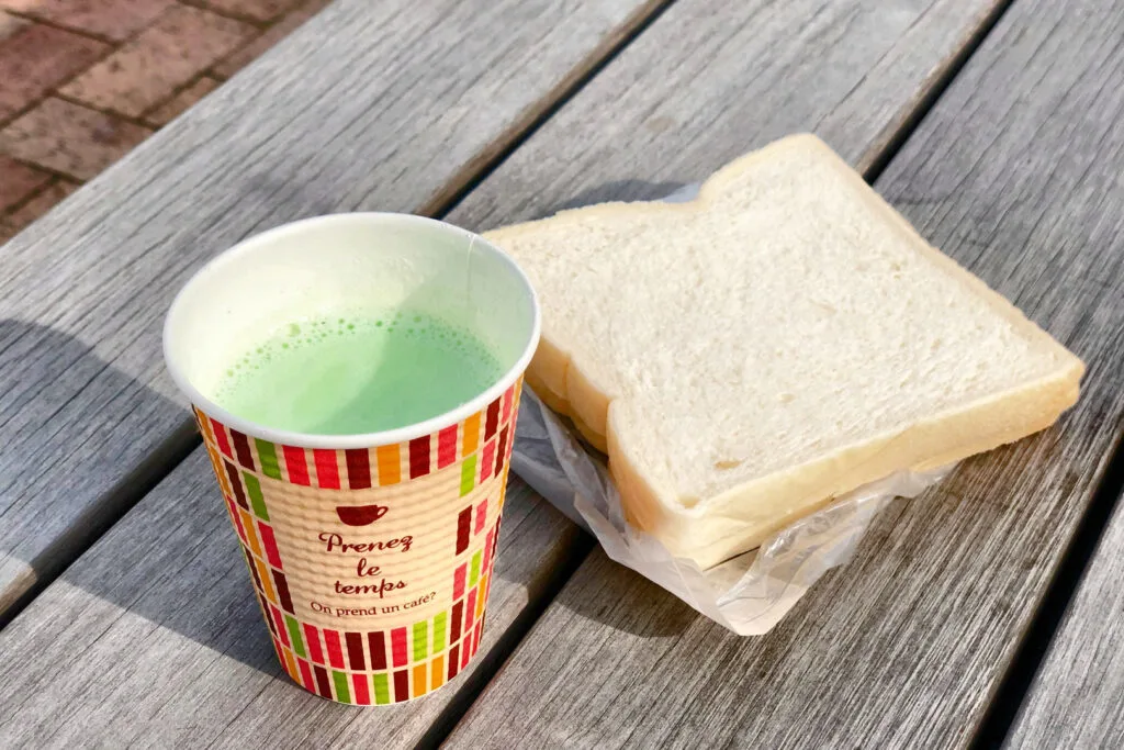 A lid-less disposable coffee cup filled with light green zunda milk and to the right of it two pieces of white bread sandwiched together with an unseen peanut filling sitting on its plastic wrapper on a park bench.