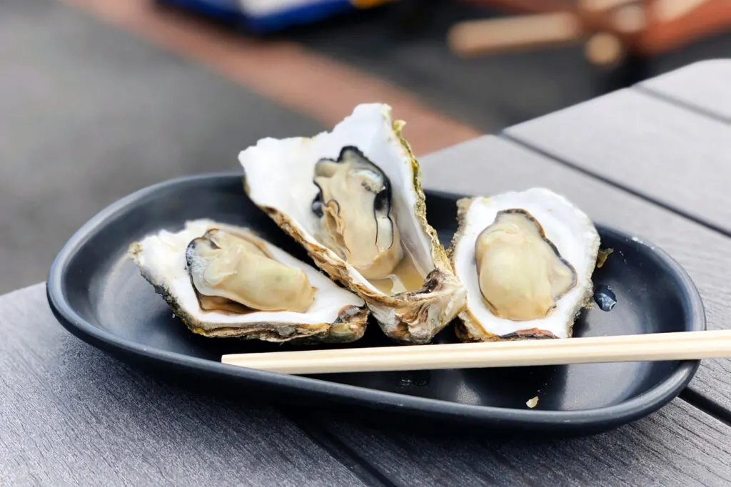 Three fresh oysters in shells on a black plate with disposable wooden chopsticks on a black wooden outdoor table.