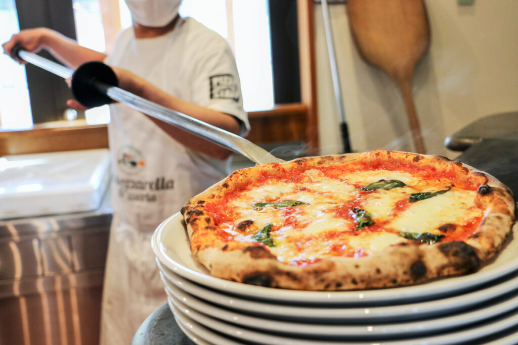 A staff member dressed in white and a white apron has just removed a margherita pizza from the oven using a large long-sticked pizza peel and is placing it on the top plate of a stack.