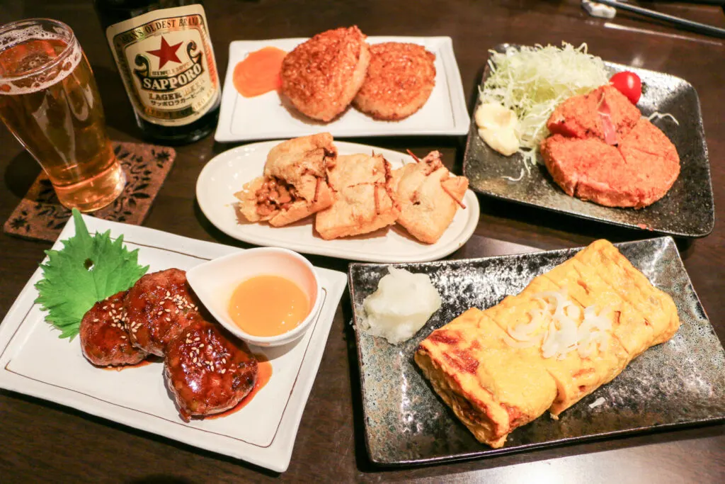 A table full of smaller dishes for sharing izakaya-style, including natto inari-age (one of the tofu pockets has been opened to show the natto filling) and sanma meat balls. A glass of beer and a beer bottle can be seen at the top-left of the image.
