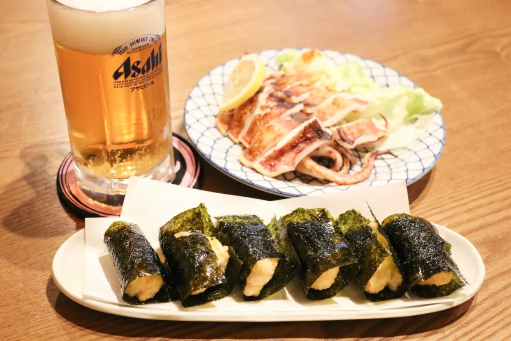 A plate of deep-fried Japanese yam dumplings rolled in nori seaweed. Behind it on the left is a glass of beer and to the right a plate of grilled semi-dried squid.