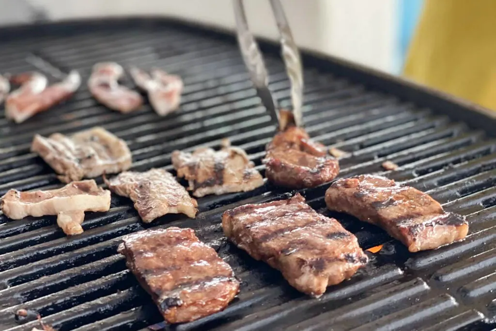 Cuts of meat being cooked on the BBQ grill. A pair of metal tongs can be seen coming in from the top-middle of the image, turning a piece of meat.
