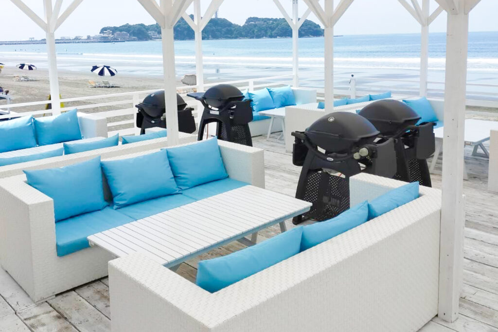 A classy tebura BBQ shack at a beach with portable BBQ grills, and white couches with light blue cushions that blend in seamlessly with the ocean in view behind.
