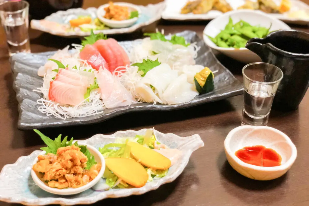 A table full of Japanese dishes including a platter of sashimi, a bowl of edamame and two glasses of sake.