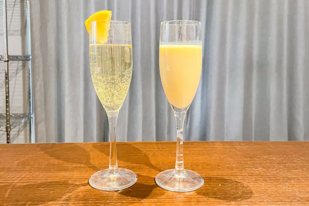 Two drinks in champagne flutes on a wooden table. The one on the left is a sparkling golden-colored cocktail with a slice of lemon on the rim of the glass. The one on the right is a banana-colored milky cocktail.