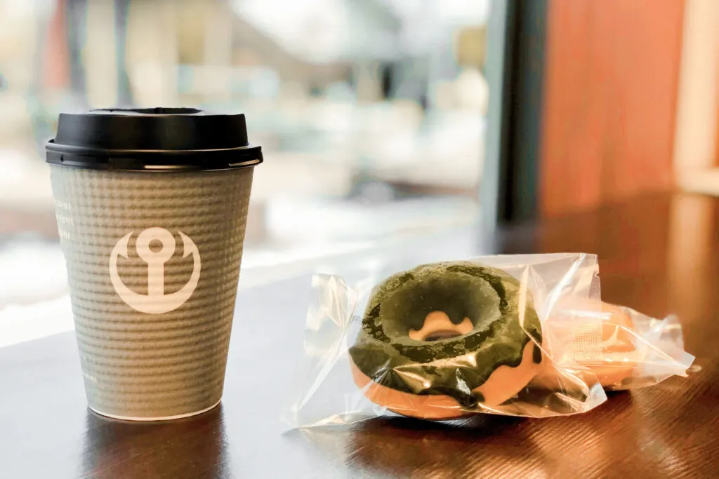 A light olive green disposable coffee cup with the company's logo in white on it and a black plastic lid. Next to it are two donuts in plastic packaging, the top one with a green-colored glaze. They sit upon a wooden window-facing countertop in the shop.